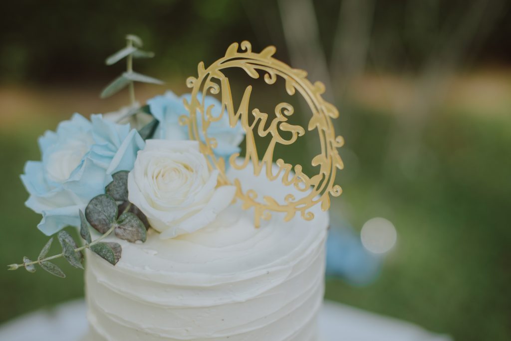 Elegant white wedding cake with gold sign on top, light blue and white flowers, and eucalyptus branches