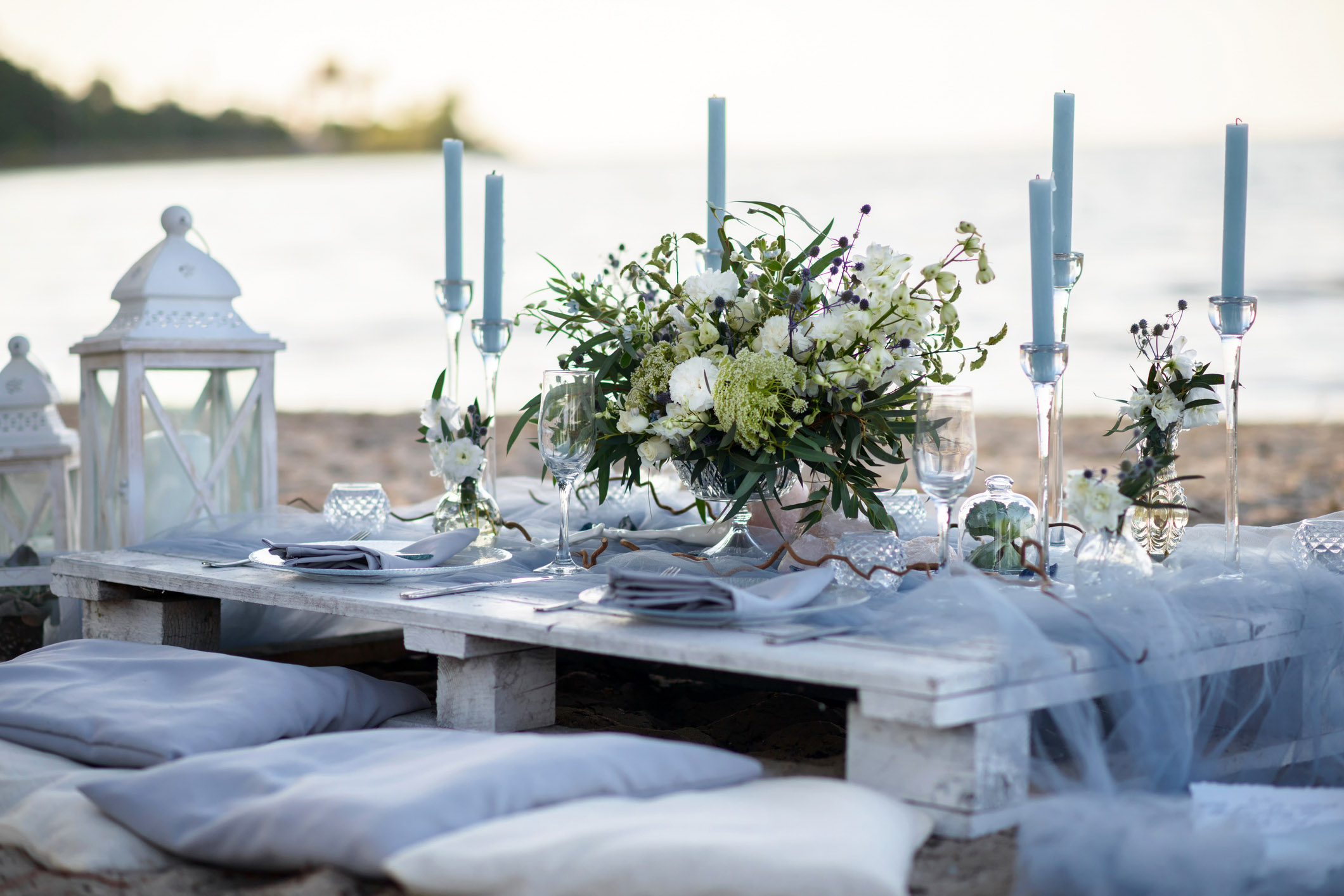 Romantic beach wedding dinner table setup for two with white and pale light blue details, matching pillows, and a centerpiece with white flowers and branches, facing the sea