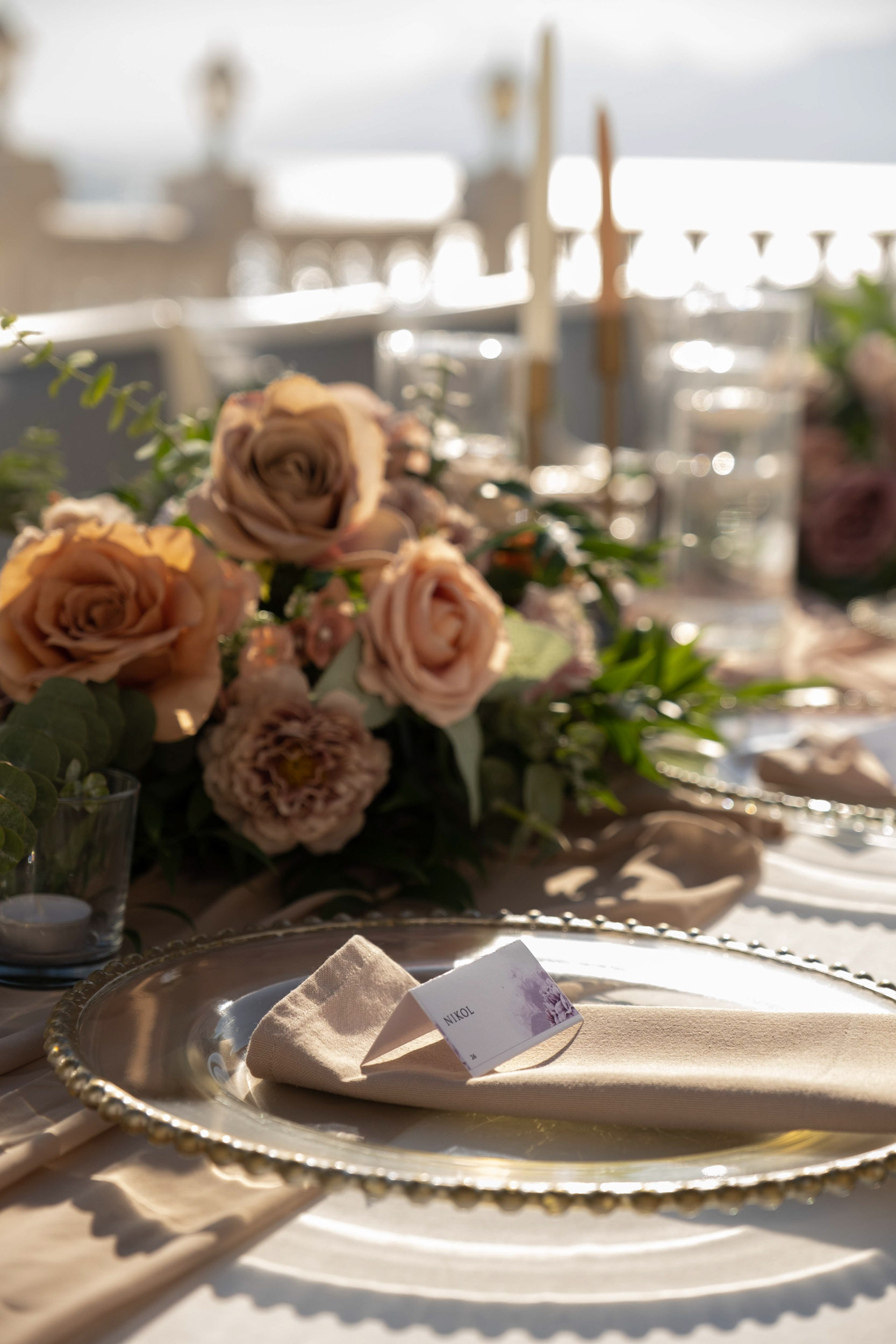 Close-up photo of a rehearsal dinner table setup with flower arrangements, gold-detailed glassware, charging plates, candle holders, and linen
