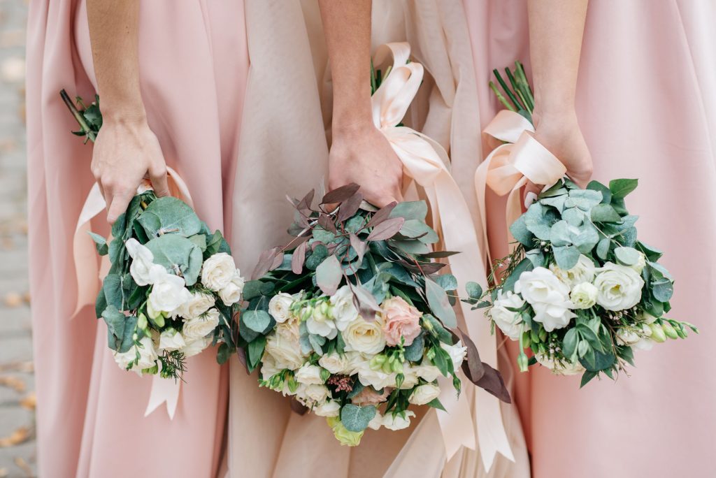 Three bridesmaids in blush pink dresses holding bouquets of white and blush pink roses, eucalyptus, and light pink satin ribbons